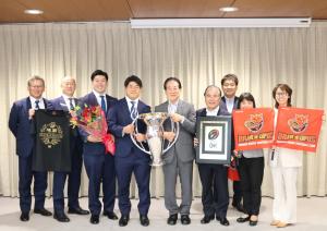 画像：市長から贈呈された花束や優勝トロフィーなどを掲げた出席者の集合写真（拡大画像へのリンク）