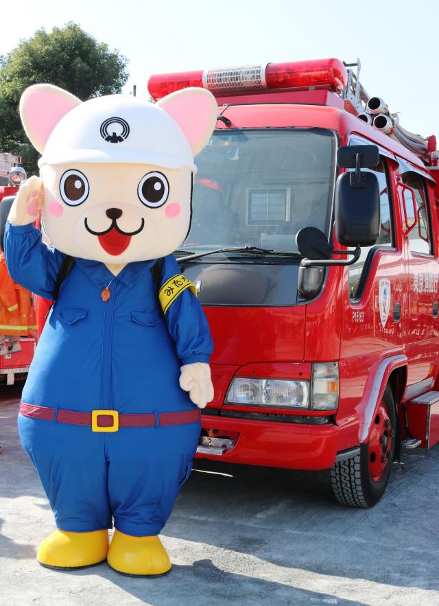 画像：写真：消防車の前で敬礼するじじょまるの着ぐるみの写真